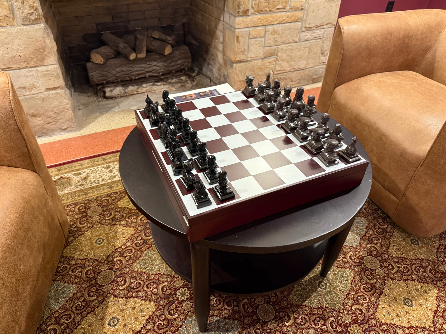 Chessboard with pieces on a round table in a cozy room with a fireplace and brown chairs.